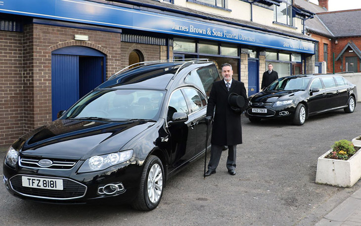 A man dressed in black standing next to a hearse in front of James Brown & Sons