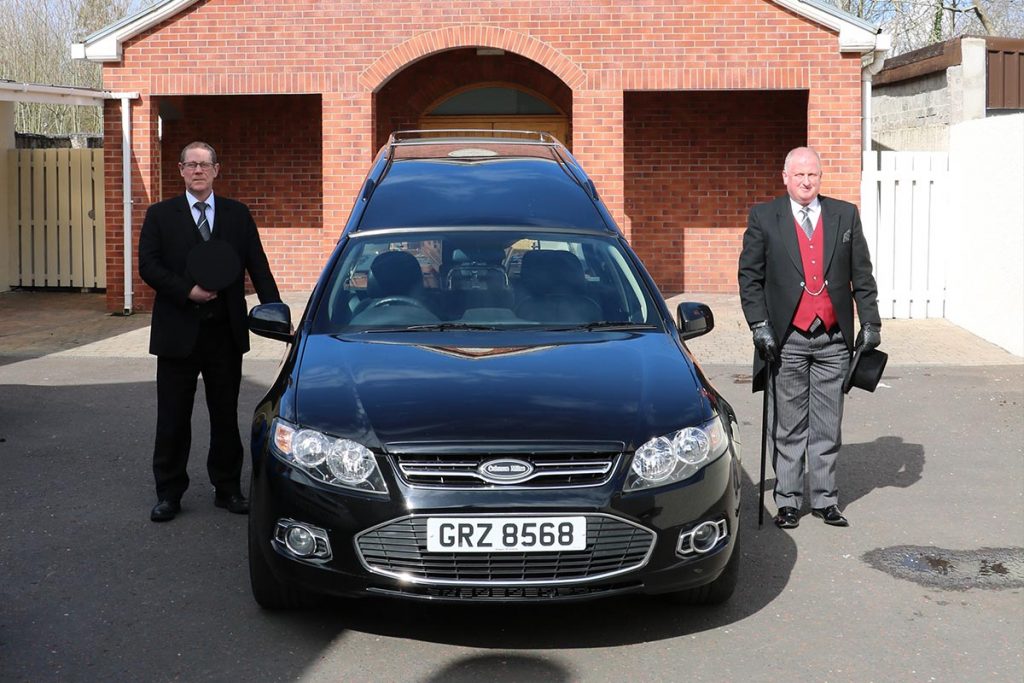 Two well-dressed men standing alongside a modern hearse