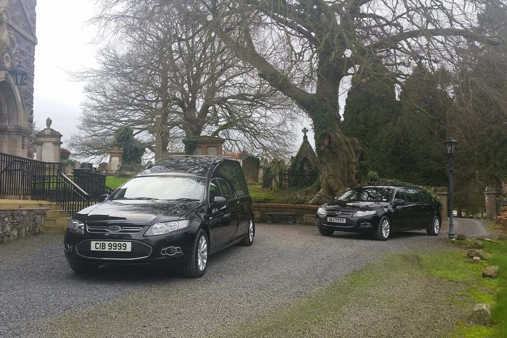 A hearse and a limousine pass by a cemetery